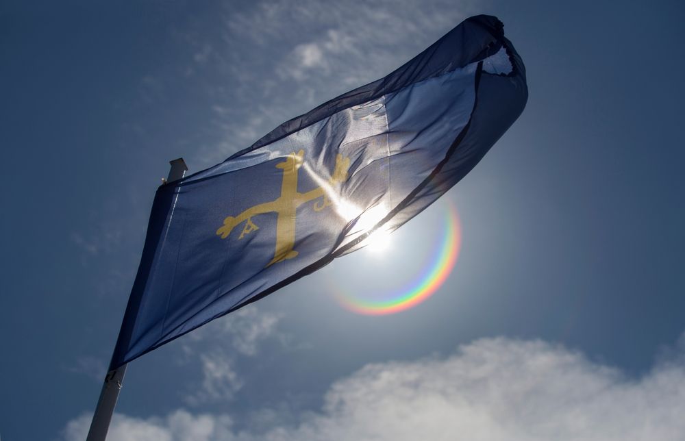 La bandera de Asturias, una cruz amarilla de la Victoria sobre fondo azul, ondea contra un cielo azul iluminado por el sol con un destello de lente.