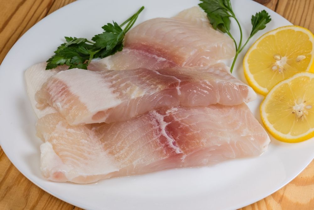 Filetes de pescado blanco crudo sobre un plato blanco, adornados con perejil fresco y dos rodajas de limón sobre una superficie de madera.