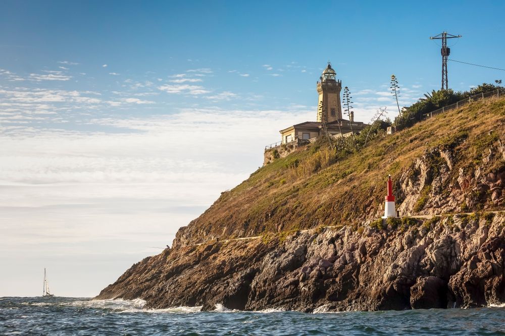 Un faro de piedra se alza sobre un acantilado costero rocoso y cubierto de hierba, con vistas al océano y bajo un cielo azul despejado.