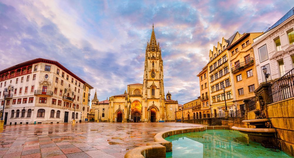 Plaza histórica europea al atardecer, con una alta iglesia gótica, suelo de piedra mojada y una fuente en primer plano.
