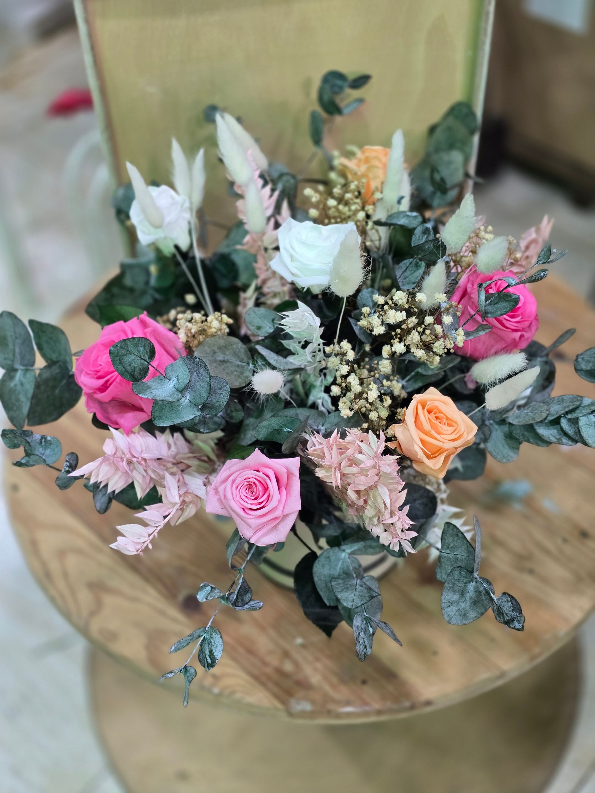 Ramo de rosas rosas, naranjas y blancas con vegetación, sentado sobre un taburete de madera.