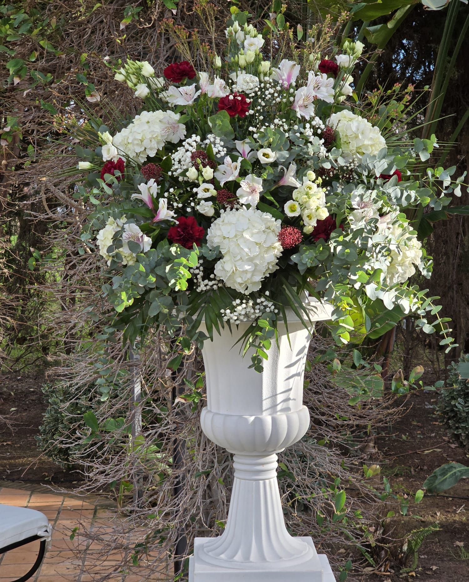 Arreglo floral blanco en una urna blanca. Incluye hortensias, flores rojas y follaje, en un ambiente exterior.