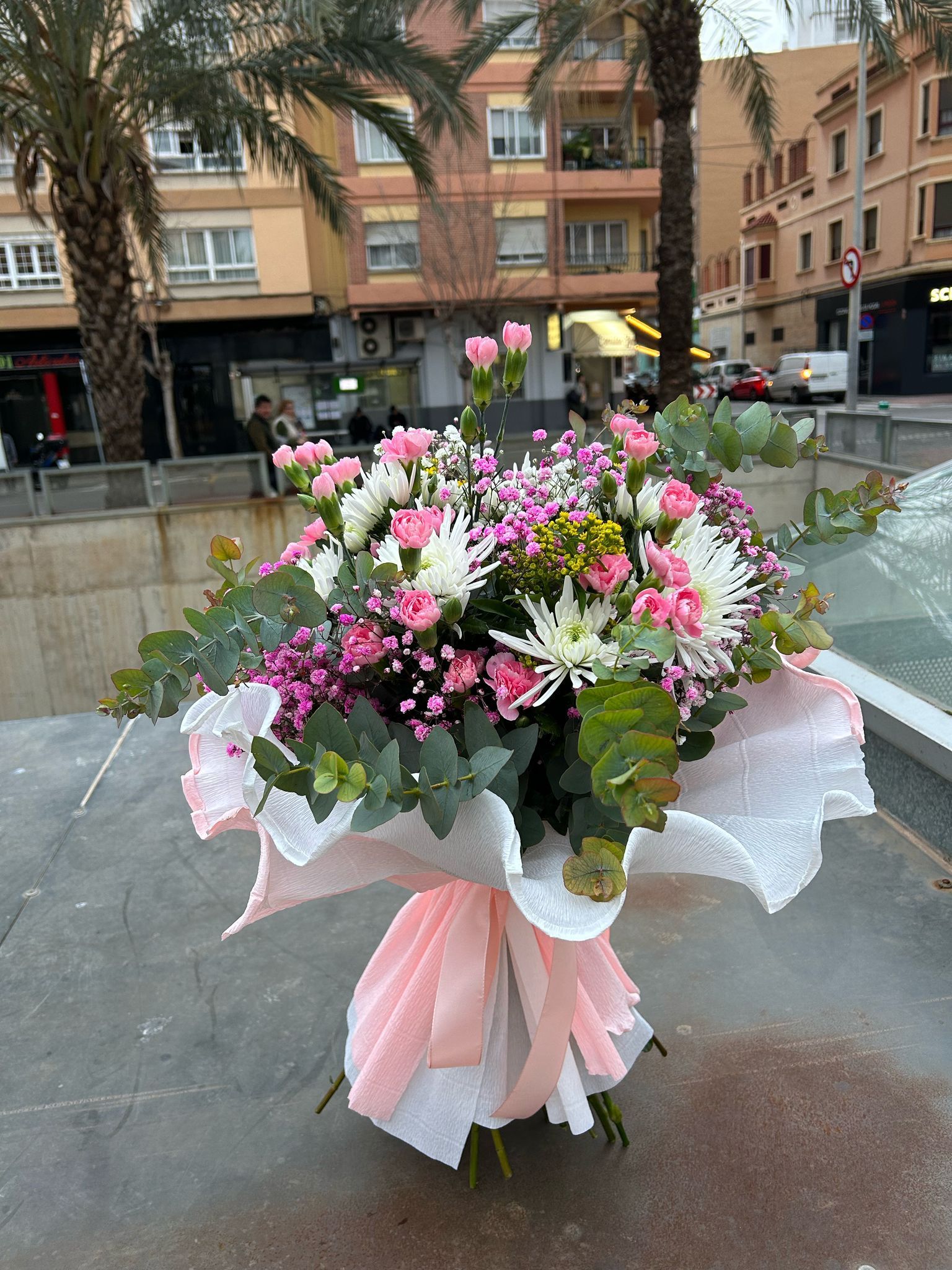 Ramo de flores rosas y blancas envuelto en papel de seda, sobre una repisa de piedra, fondo urbano.