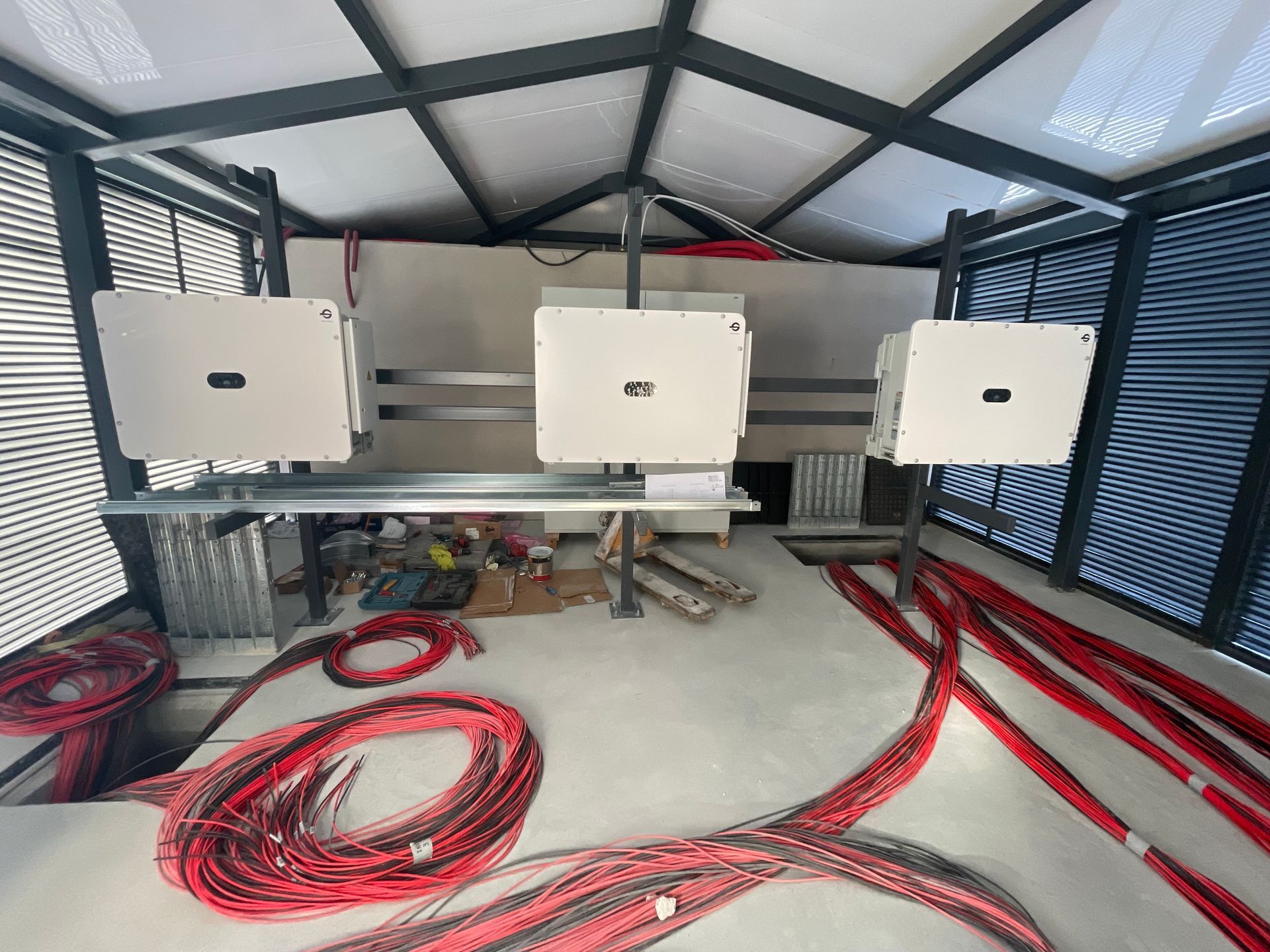 Dentro de un edificio, tres inversores solares blancos están montados en un marco de metal, con cables rojos en el piso.