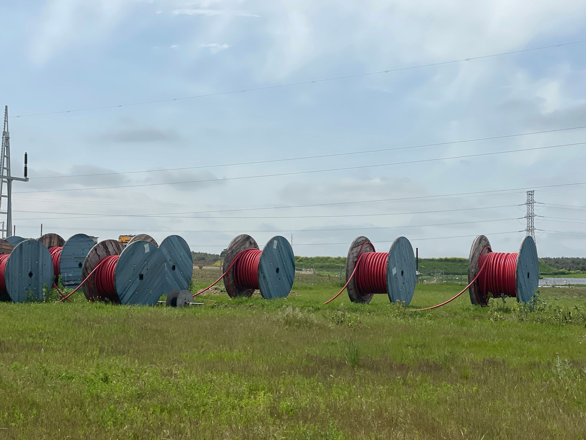Carrete de cable rojo y gris reposa sobre un campo de hierba con una torre de energía al fondo bajo un cielo nublado.