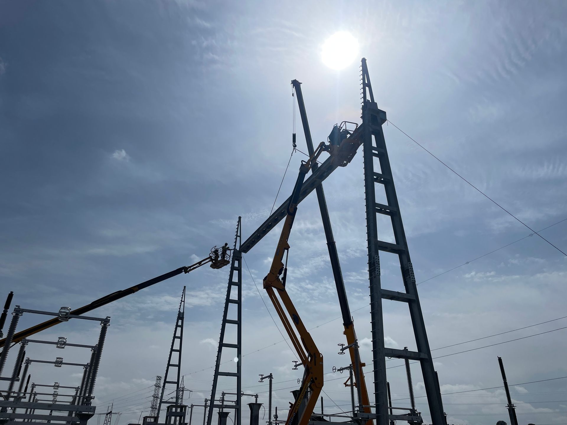 Trabajadores en ascensores instalando líneas eléctricas bajo un cielo nublado. El sol brilla alto.