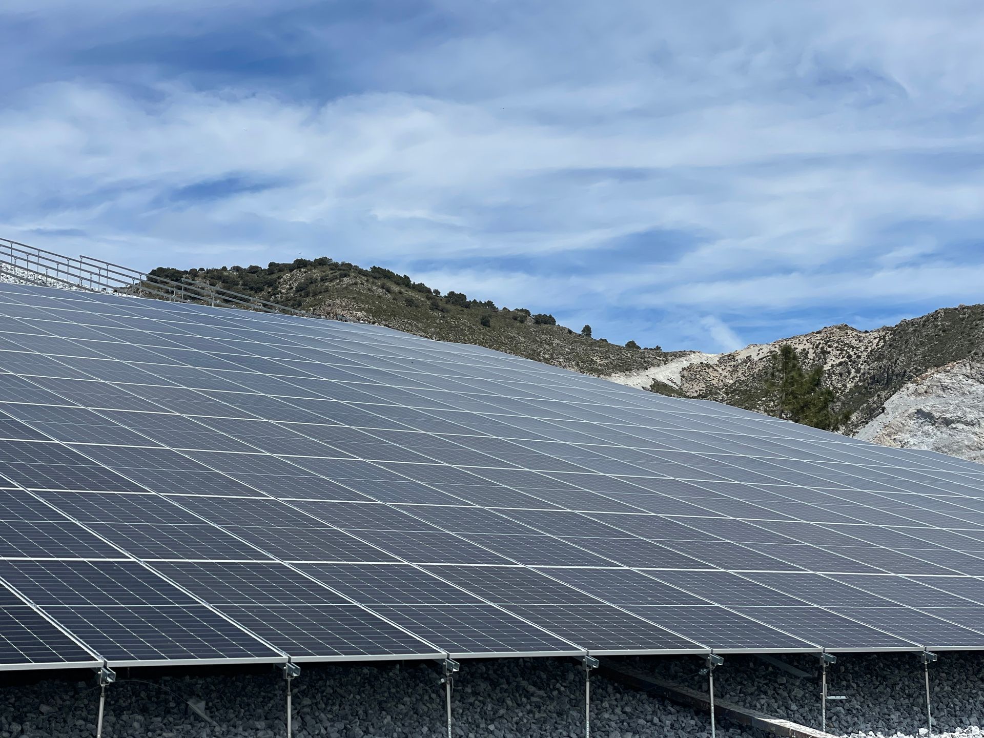 Paneles solares en una ladera con montañas al fondo bajo un cielo parcialmente nublado.