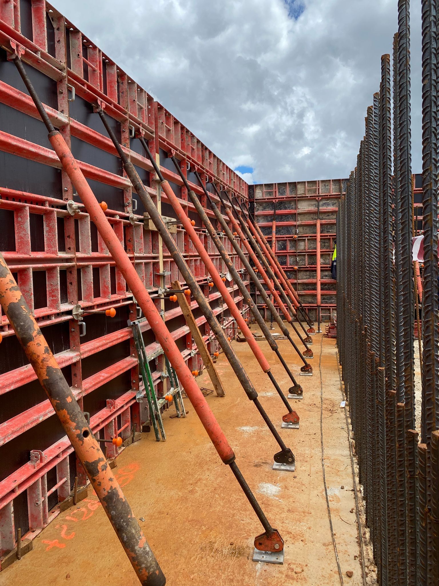 Obra: Muros de encofrado rojo sostenidos por tirantes naranjas; estructura de varillas de refuerzo; cielo nublado.