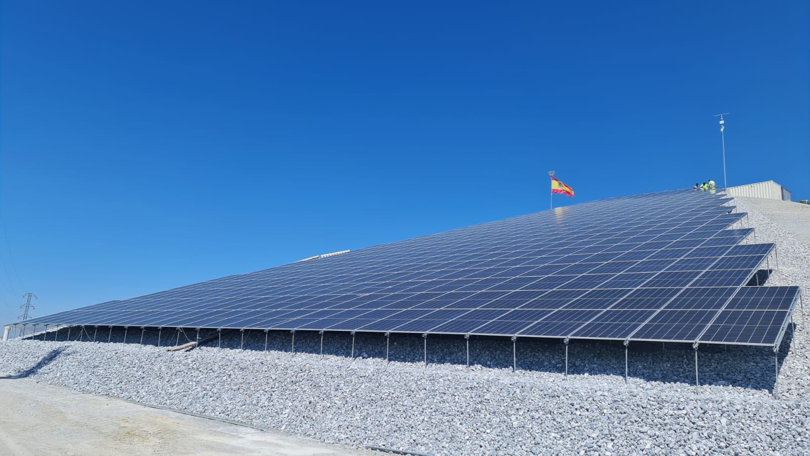 Paneles solares en una ladera bajo un cielo azul claro, con la bandera española al fondo.