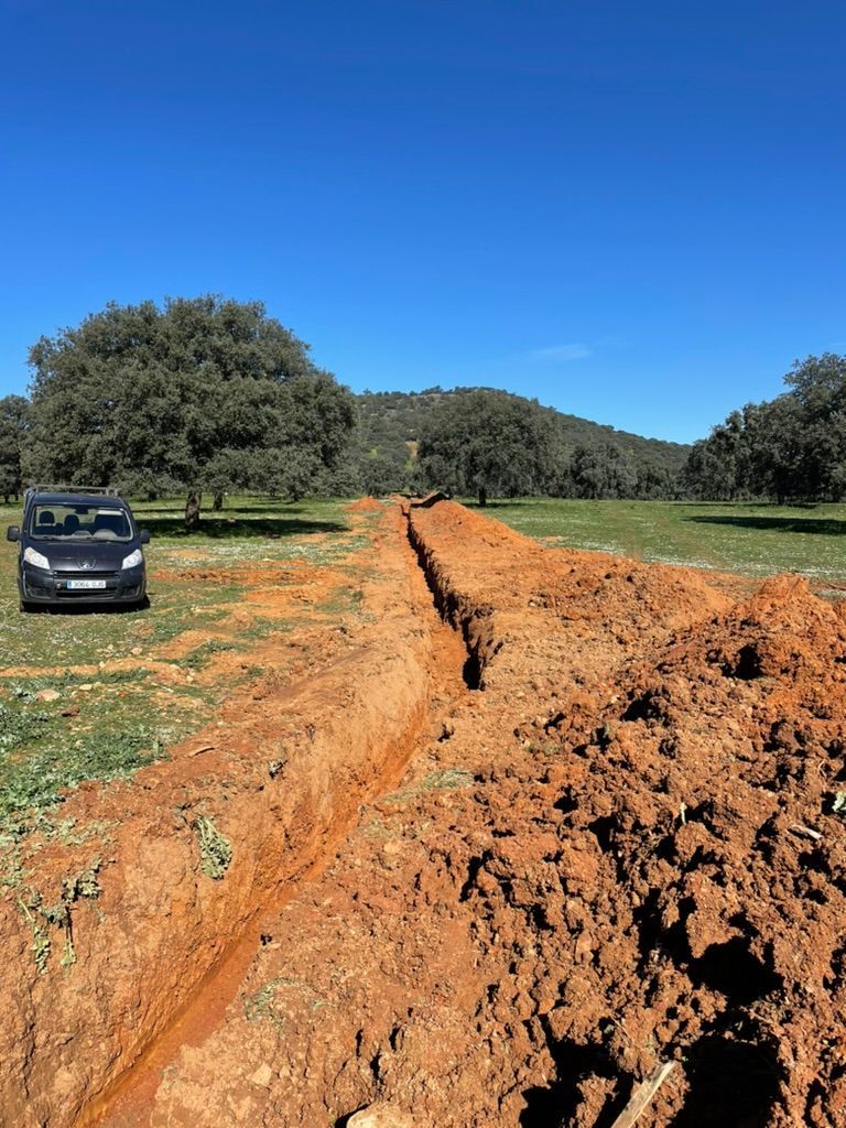Una zanja cavada en tierra roja a través de un campo de hierba, con un vehículo oscuro estacionado cerca bajo un cielo azul.