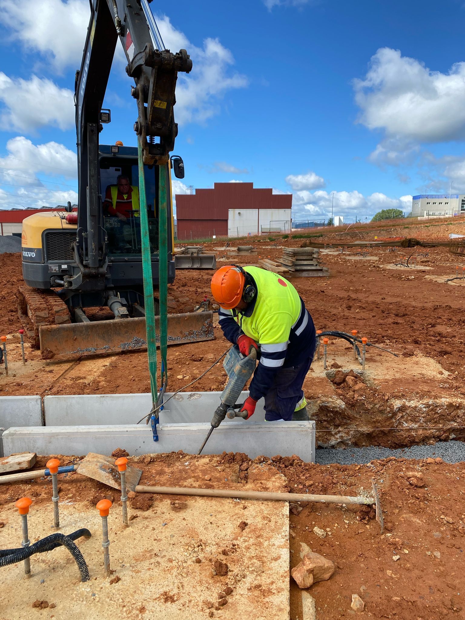 Obrero de la construcción usando un martillo neumático sobre hormigón, excavadora en lo alto, cielo azul, tierra roja.
