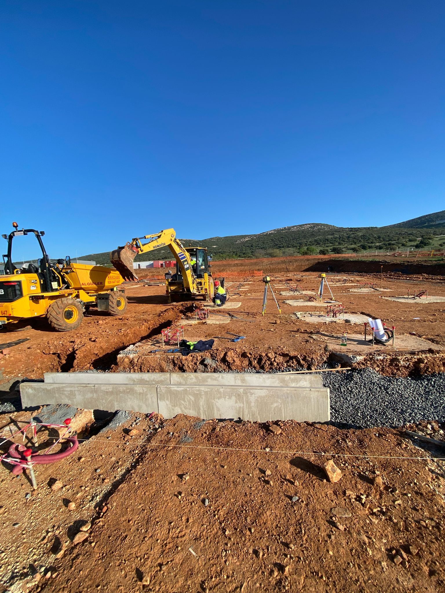 Sitio de construcción con excavadoras amarillas, trabajadores y cielo azul claro.