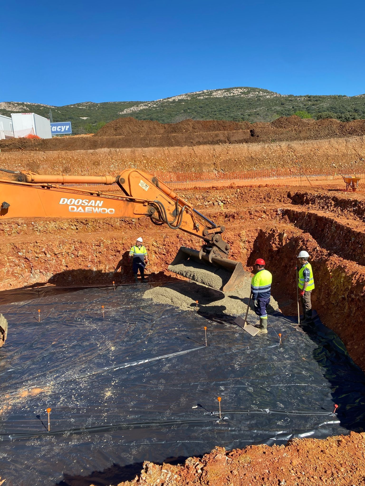 Obra: excavadora, trabajadores y revestimiento negro en un pozo. Día soleado, tierra roja, cielo azul.