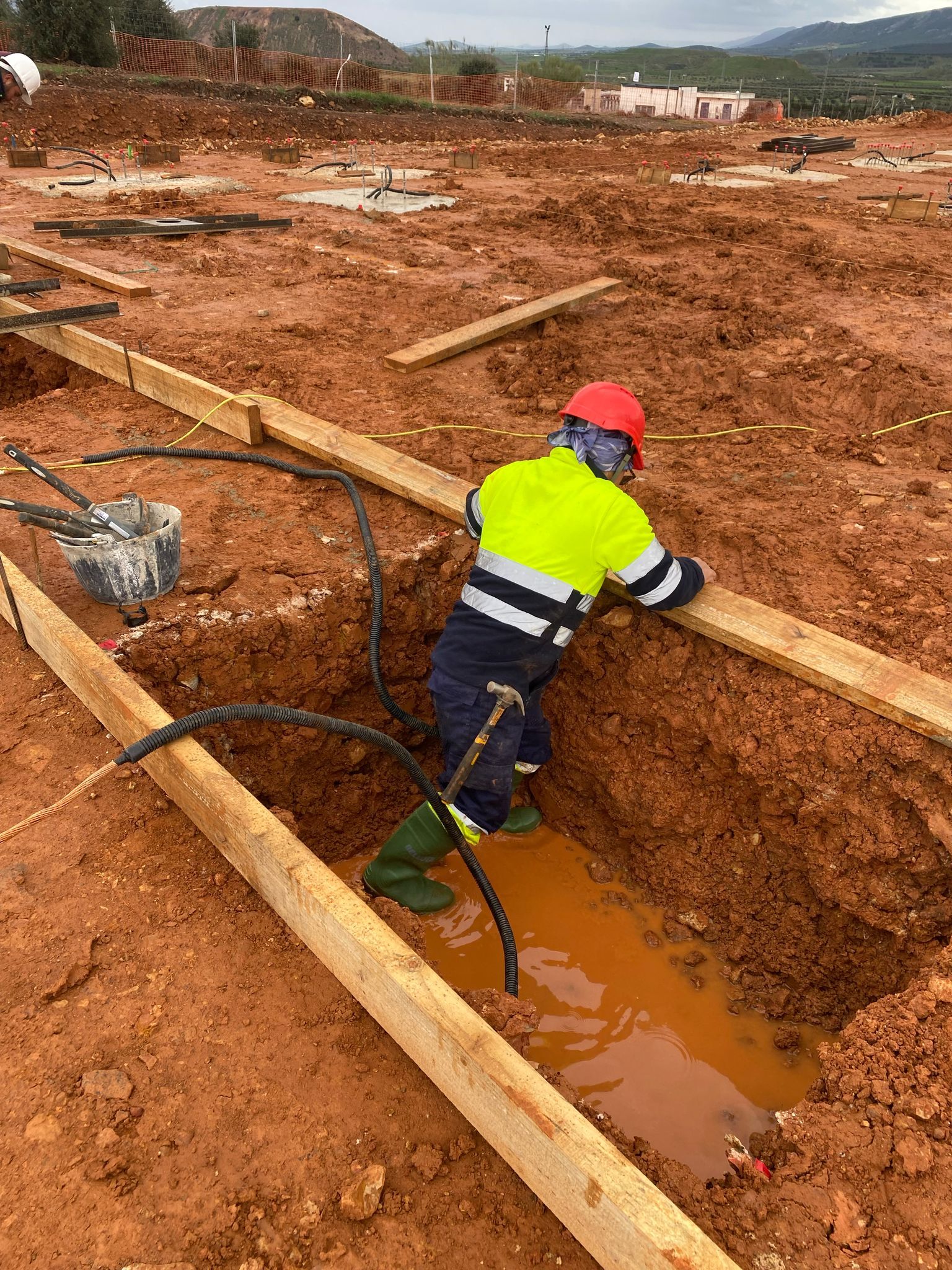 Obrero en una zanja lodosa instalando cables. Sombrero rojo, chaleco amarillo, tierra y tablones de madera.
