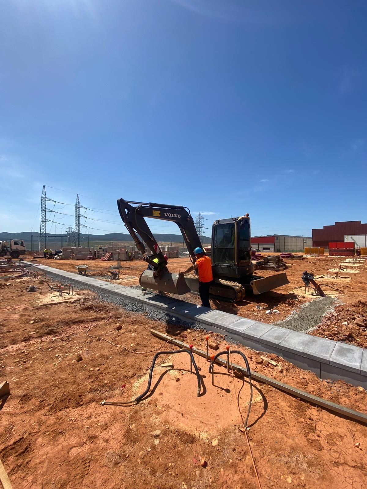 Un obrero opera una excavadora cerca de un borde pavimentado en un día soleado. Cables eléctricos y edificios al fondo.