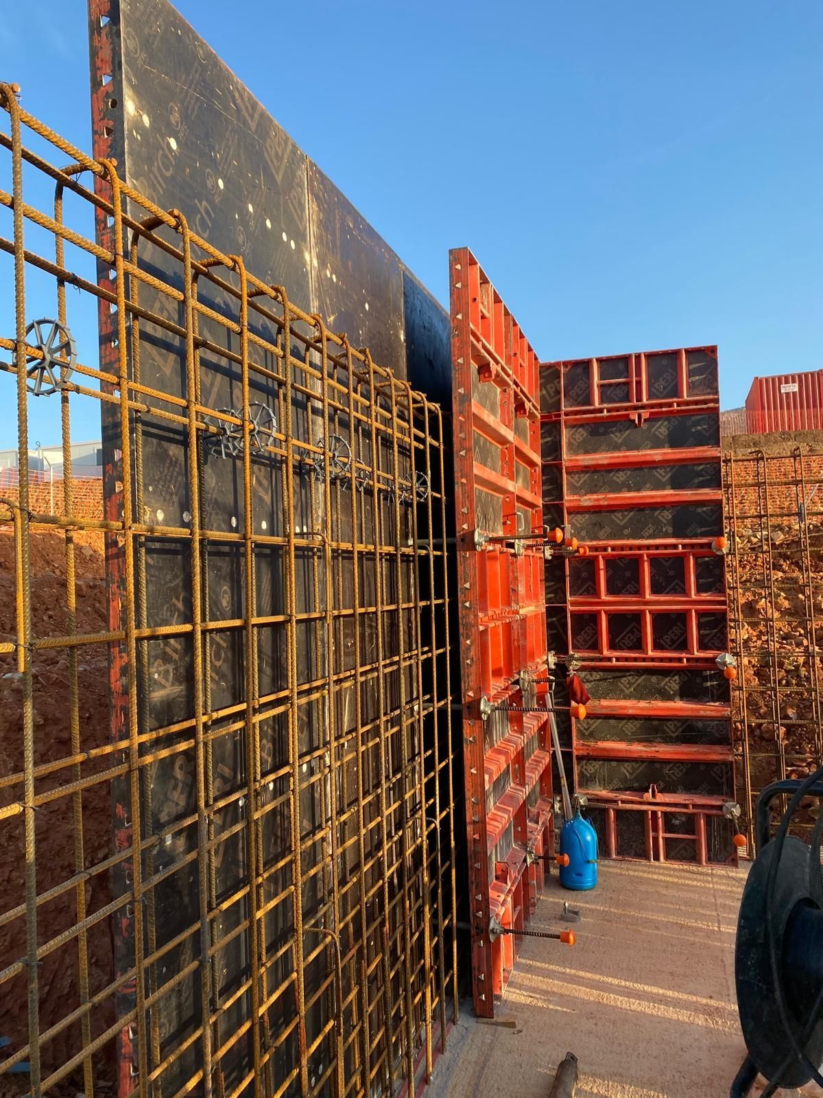 Obra con varillas corrugadas y encofrado de hormigón. Paneles negros y naranjas con tanque azul, de día.