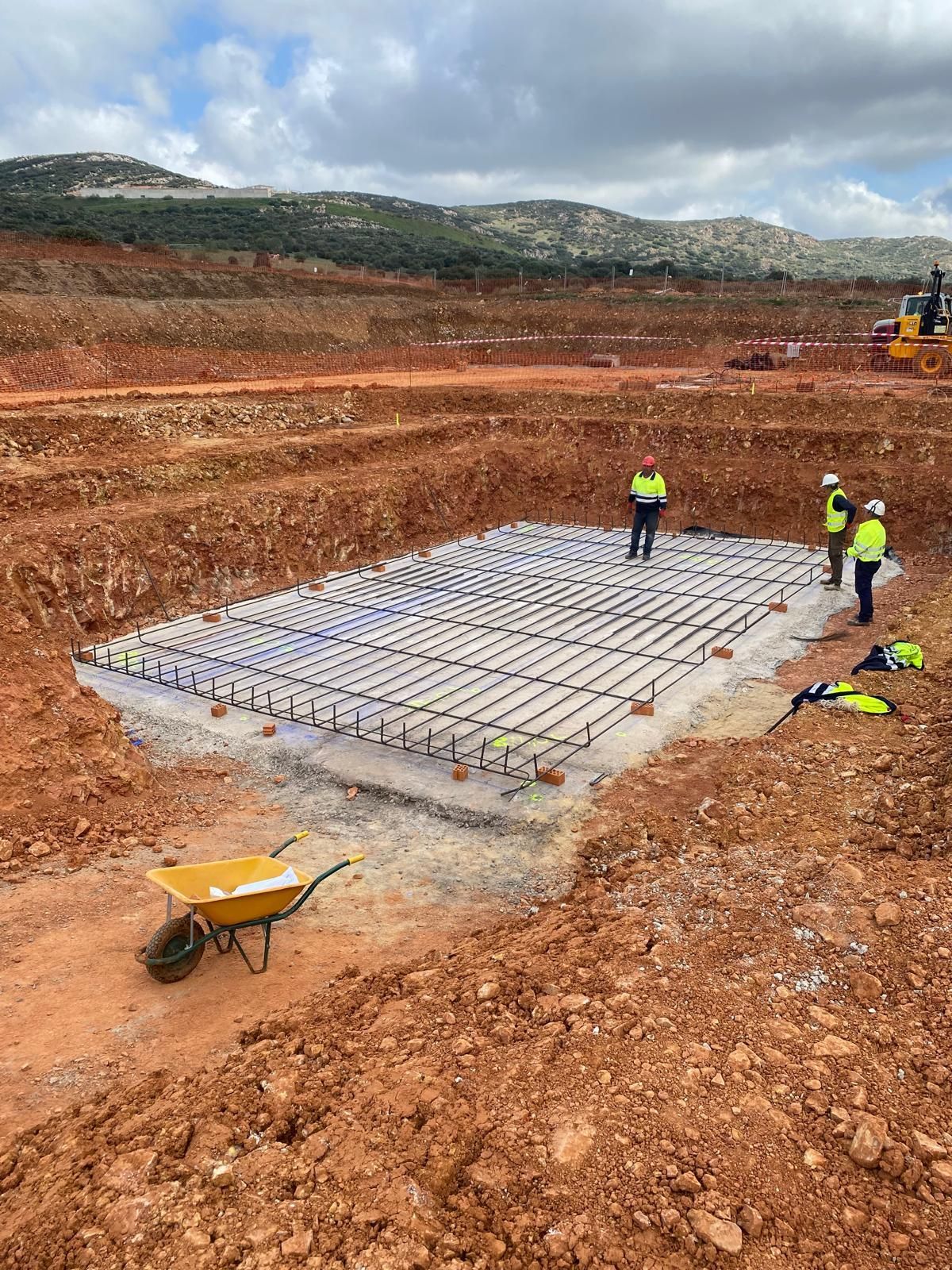 Obra: trabajadores alrededor de una base rectangular de hormigón con barras de refuerzo, en una gran excavación.