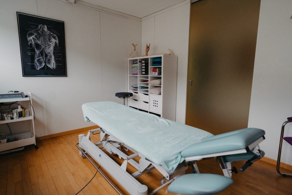 Untersuchungsraum mit hellblauer Untersuchungsliege, medizinischem Material, anatomischer Tafel und Holzboden.