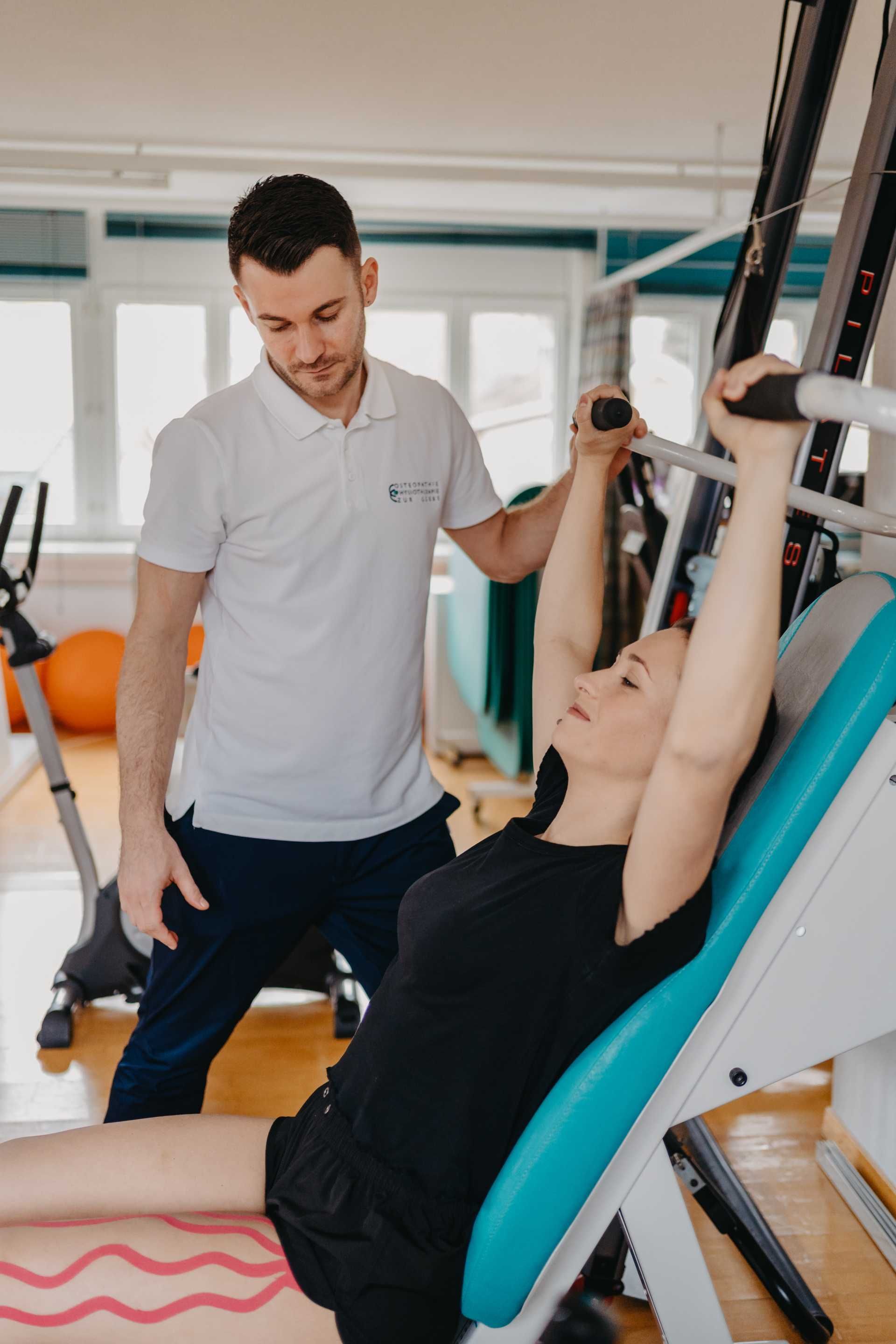 Frau trainiert mit Unterstützung eines Trainers an Fitnessgeräten. Lichtdurchfluteter Raum mit blauen und orangen Akzenten.