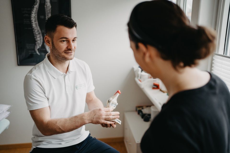 Ein Mann in einem weissen Poloshirt zeigt einer Frau in einem schwarzen Hemd in einem Innenraum ein Handmodell.