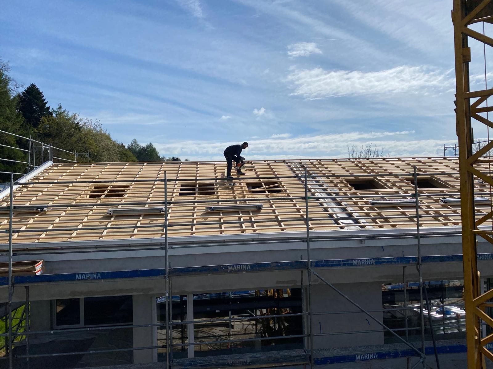 Zwei Männer stehen auf ungedecktem Dach, ein Kran hebt Palette mit Dachziegeln auf das Dach