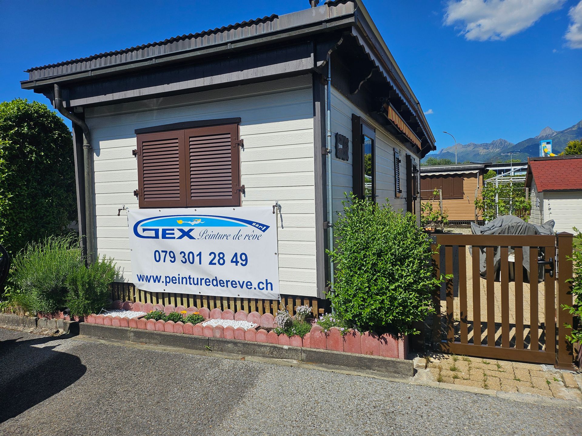 Petit bâtiment blanc avec volets et boiseries marron, panneau « Rénovation Gex », clôture en bois et buissons.