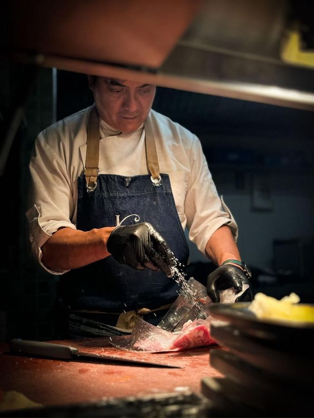 Chef en la cocina, vestido con delantal y guantes, prepara comida en una encimera roja.