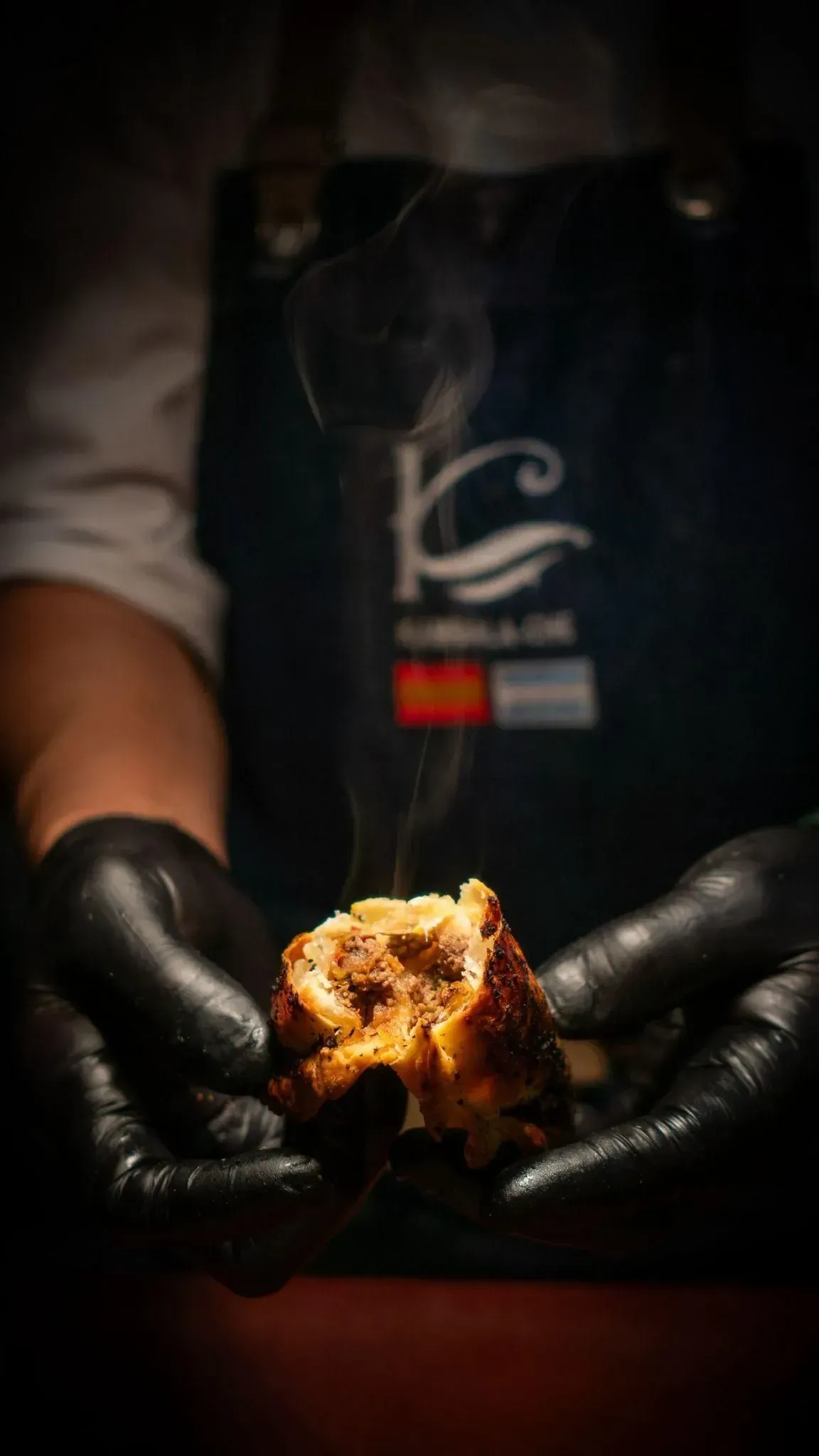 Persona con guantes negros sosteniendo un trozo humeante de comida cocinada delante de un delantal oscuro.