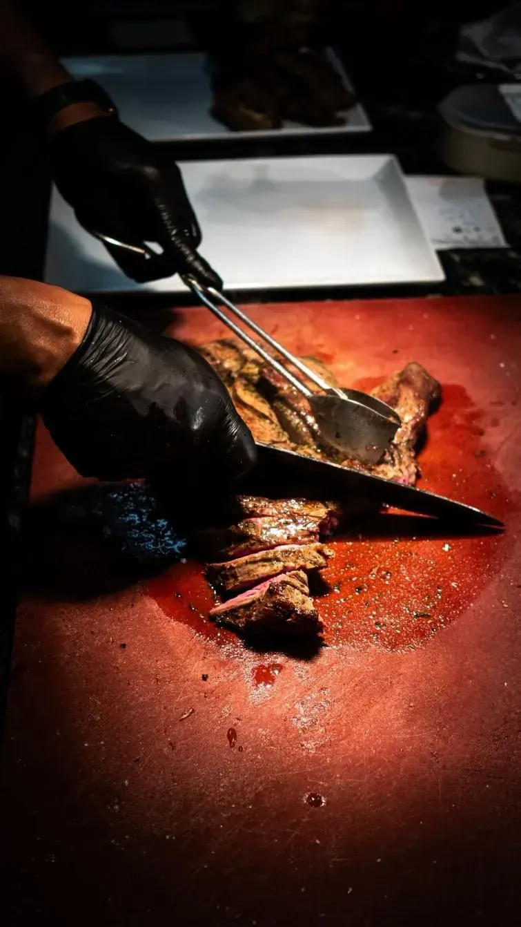 Chef con guantes negros cortando un filete cocido en una tabla de cortar roja con pinzas, cerca de platos blancos vacíos.