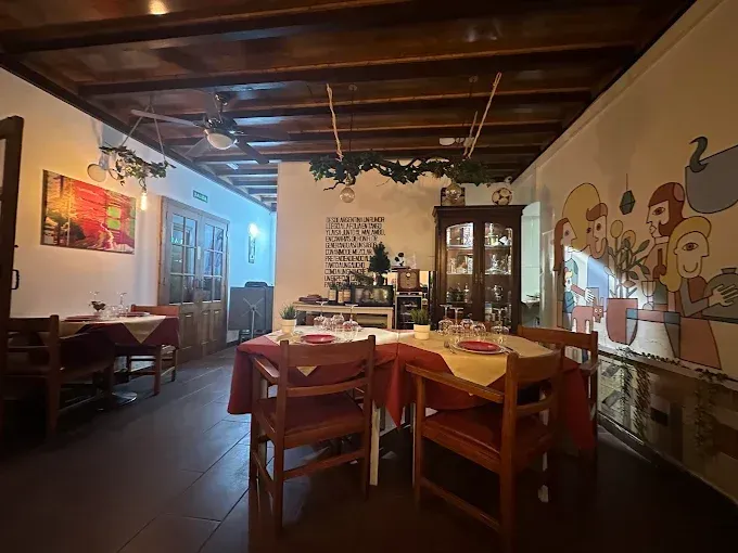 Interior del restaurante: mesas de madera con manteles rojos, mural en la pared y plantas colgantes decorativas.