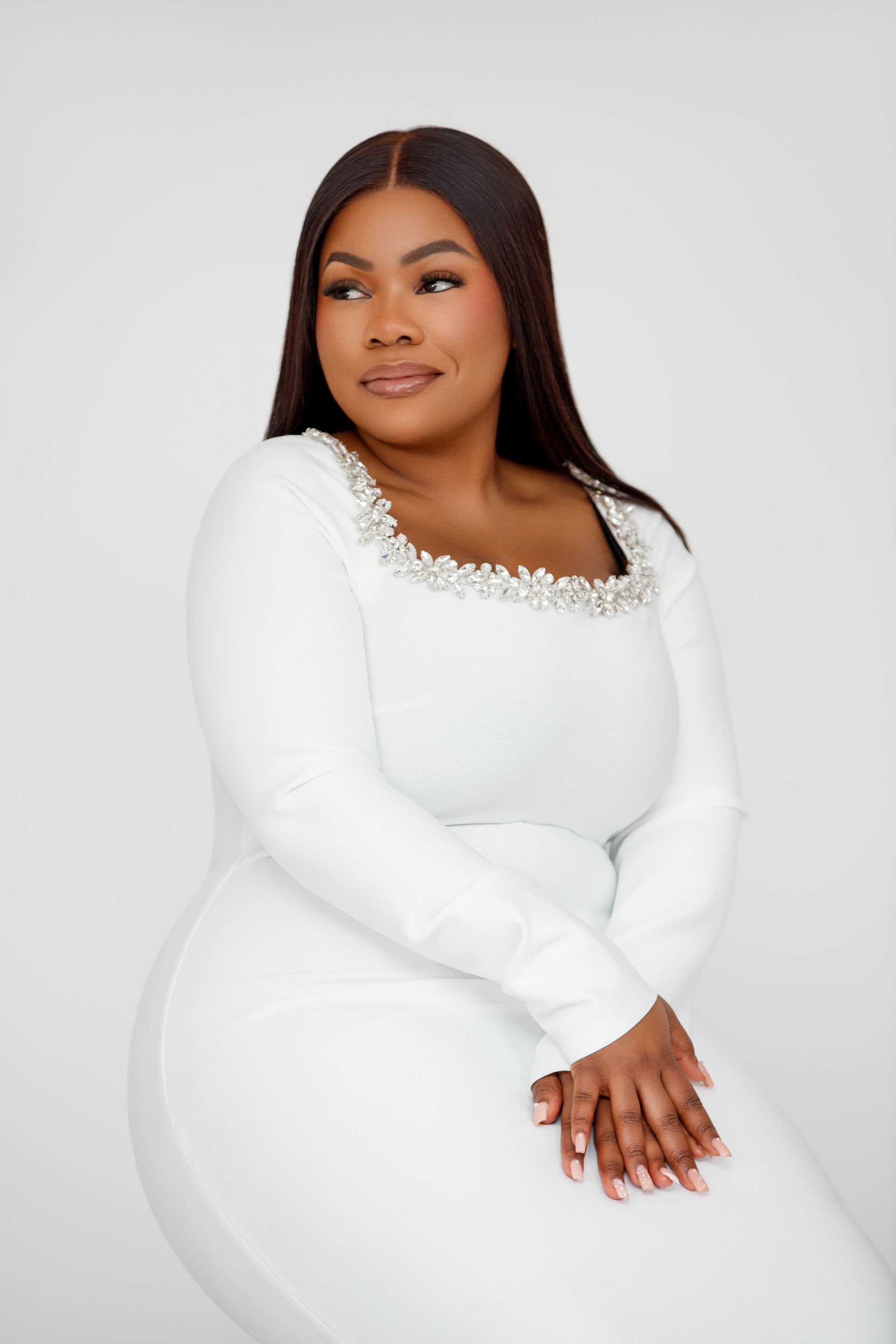 Une femme en tailleur blanc est assise, les bras croisés, sur un tabouret noir.