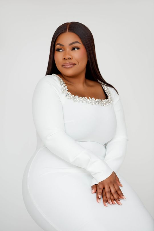 Une femme en tailleur blanc est assise, les bras croisés, sur un tabouret noir. 