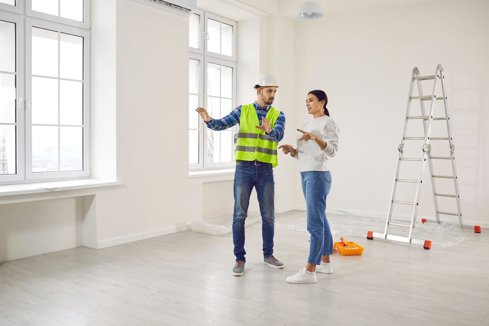 A construction professional in a vest and hard hat discusses plans with a colleague in a bright, empty room with a ladder.