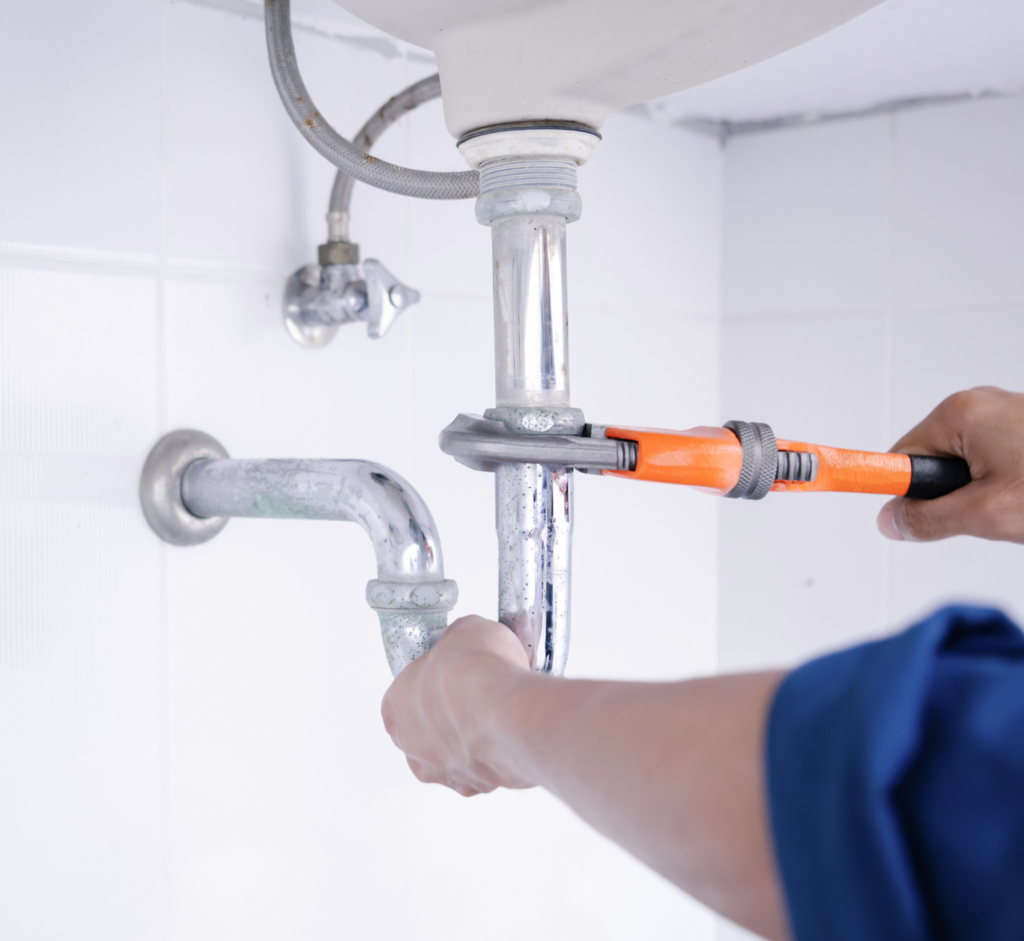 Fontanero utiliza una llave inglesa naranja para apretar las tuberías debajo de un lavabo blanco en un baño alicatado.