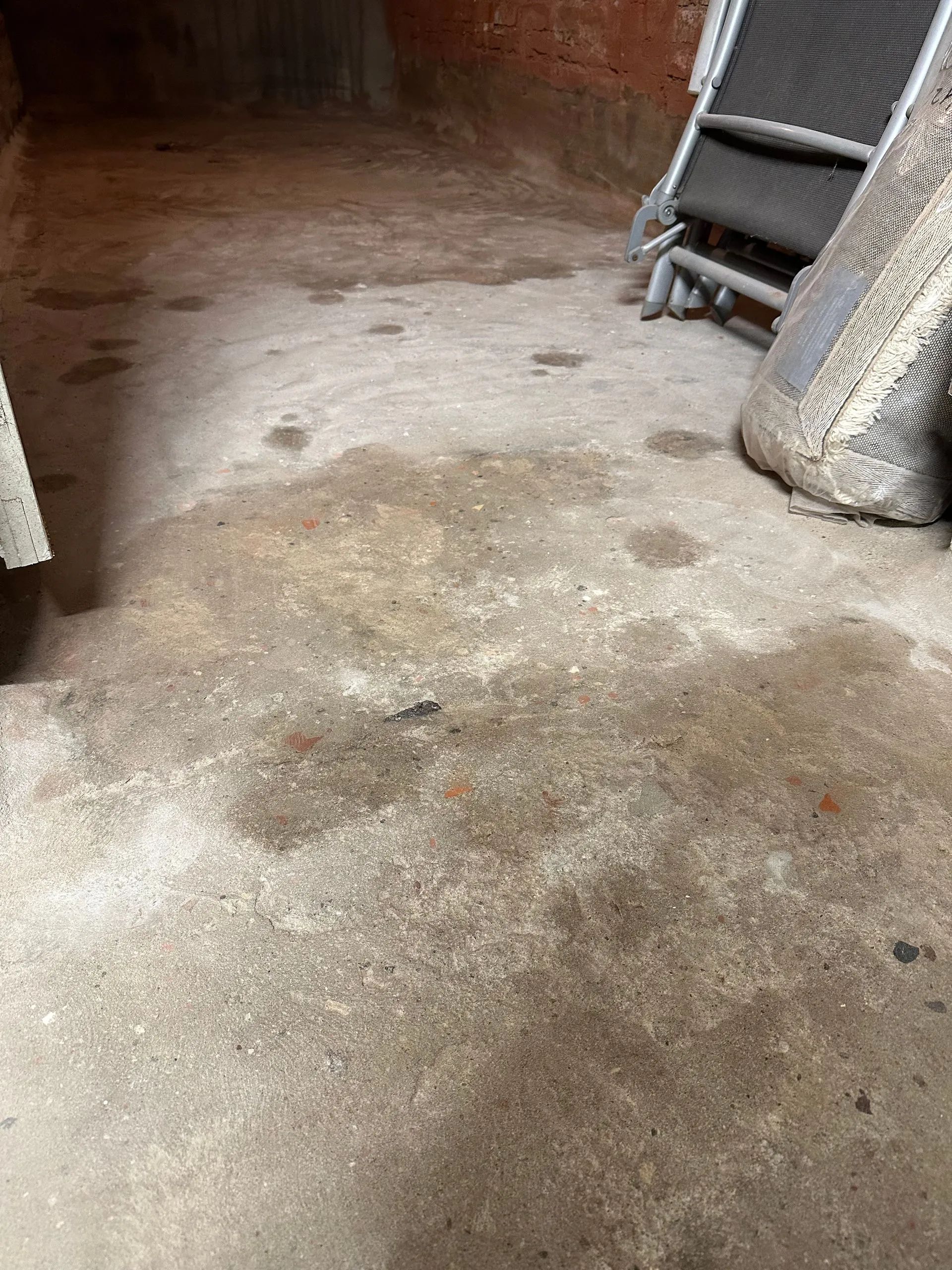 Betonboden mit Wasserflecken; im Hintergrund sind Gartenmöbel gestapelt.