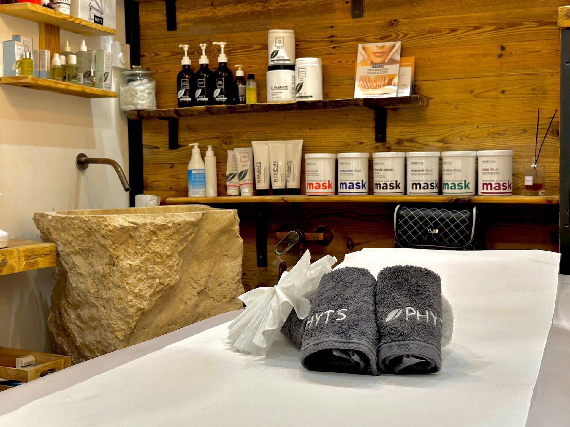 Salle de soins avec étagères en bois, produits et serviettes grises roulées sur une table de massage.