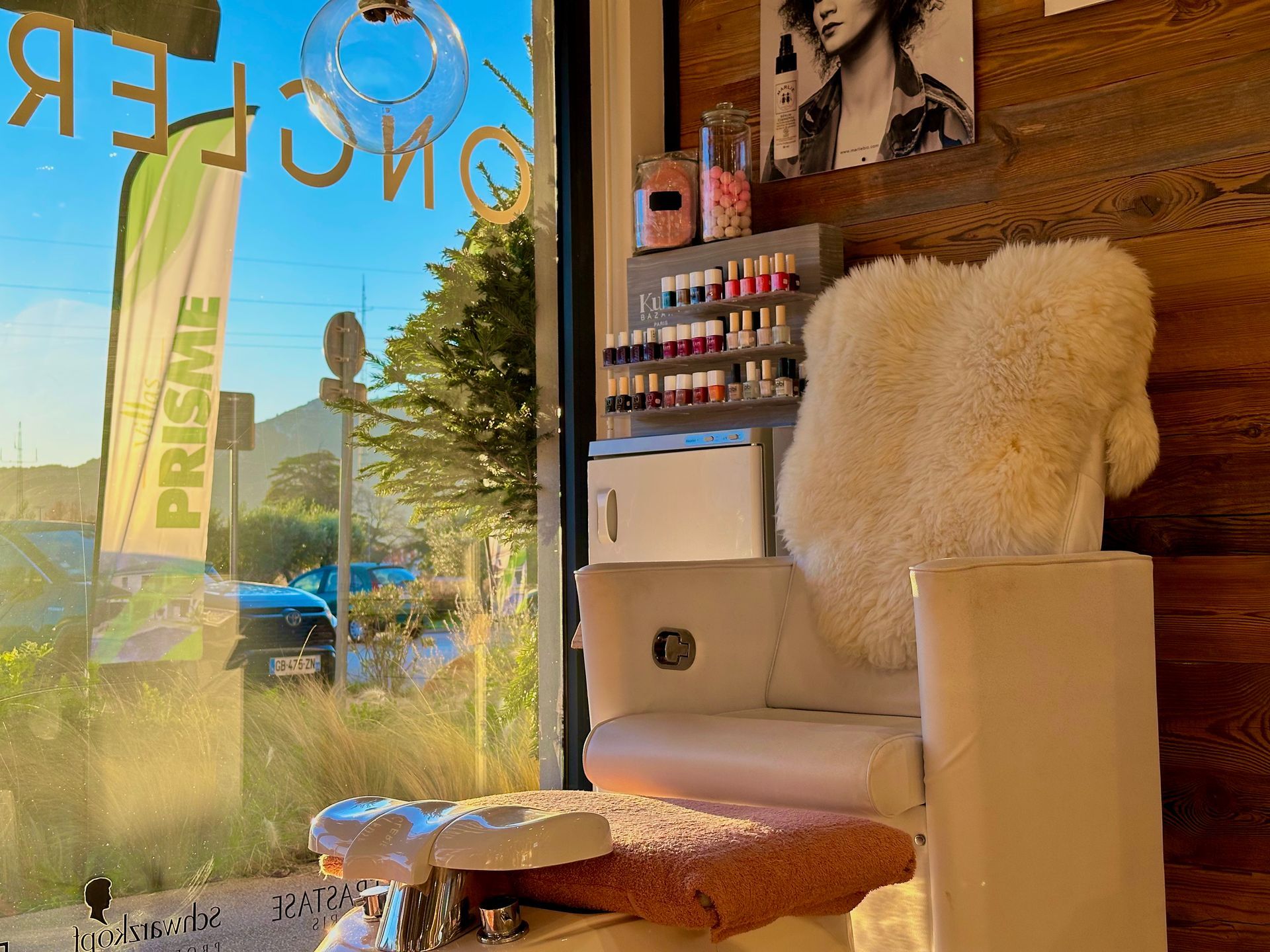 Salle d'attente cosy avec fauteuil blanc, étagères de vernis à ongles et vitrine ensoleillée.