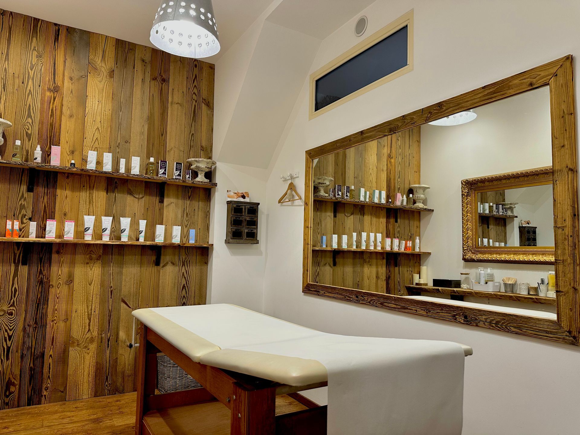 Salle de soins avec étagères en bois remplies de flacons, table de massage et grands miroirs encadrés.