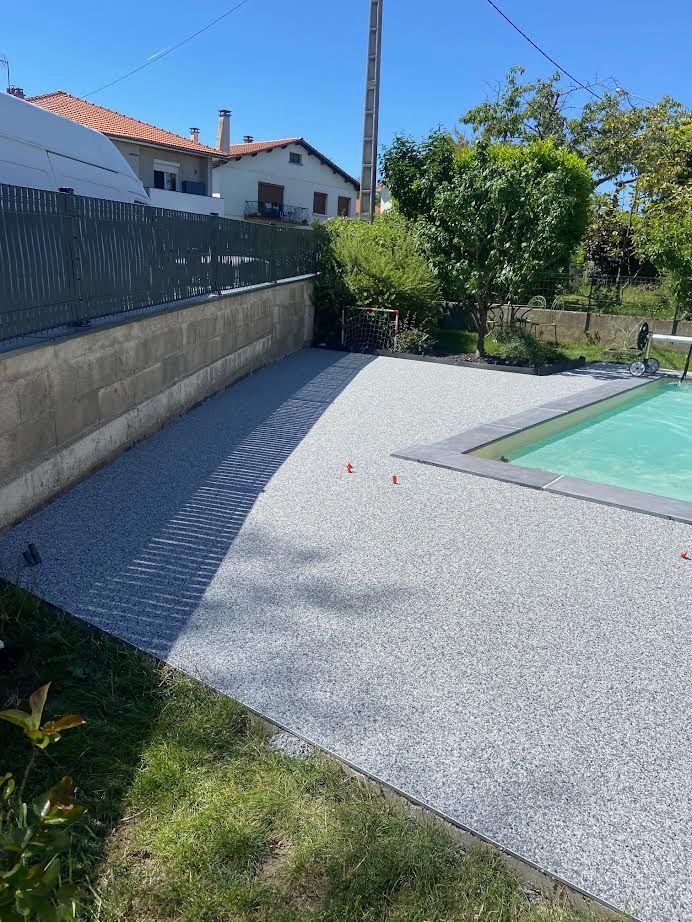 Terrasse arrière avec allée de gravier, pelouse verte, arbres, mur de pierre et bord de piscine sous un ciel bleu