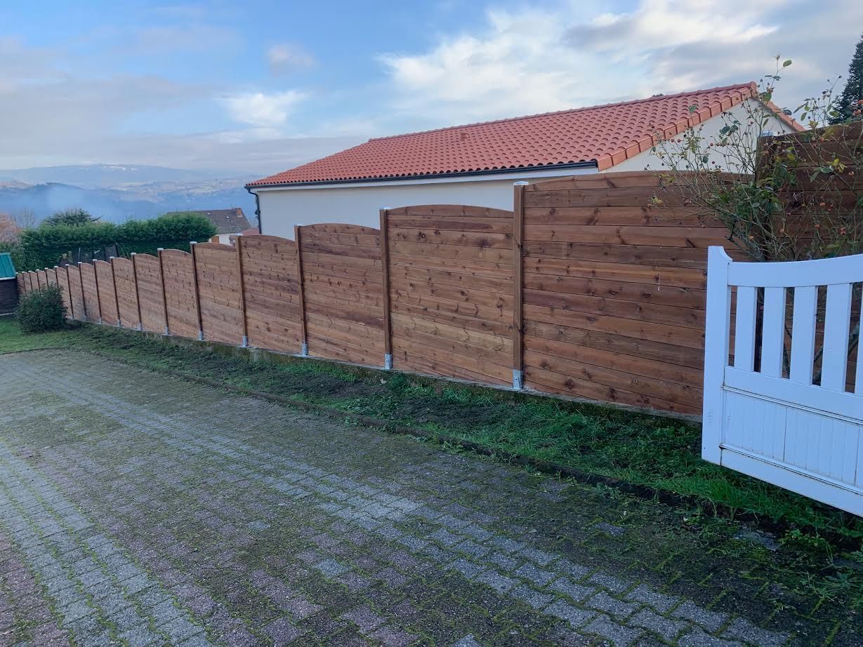Clôture en bois le long d'un chemin de gravier, à côté d'un portail blanc et d'une maison au toit rouge sous un ciel nuageux.