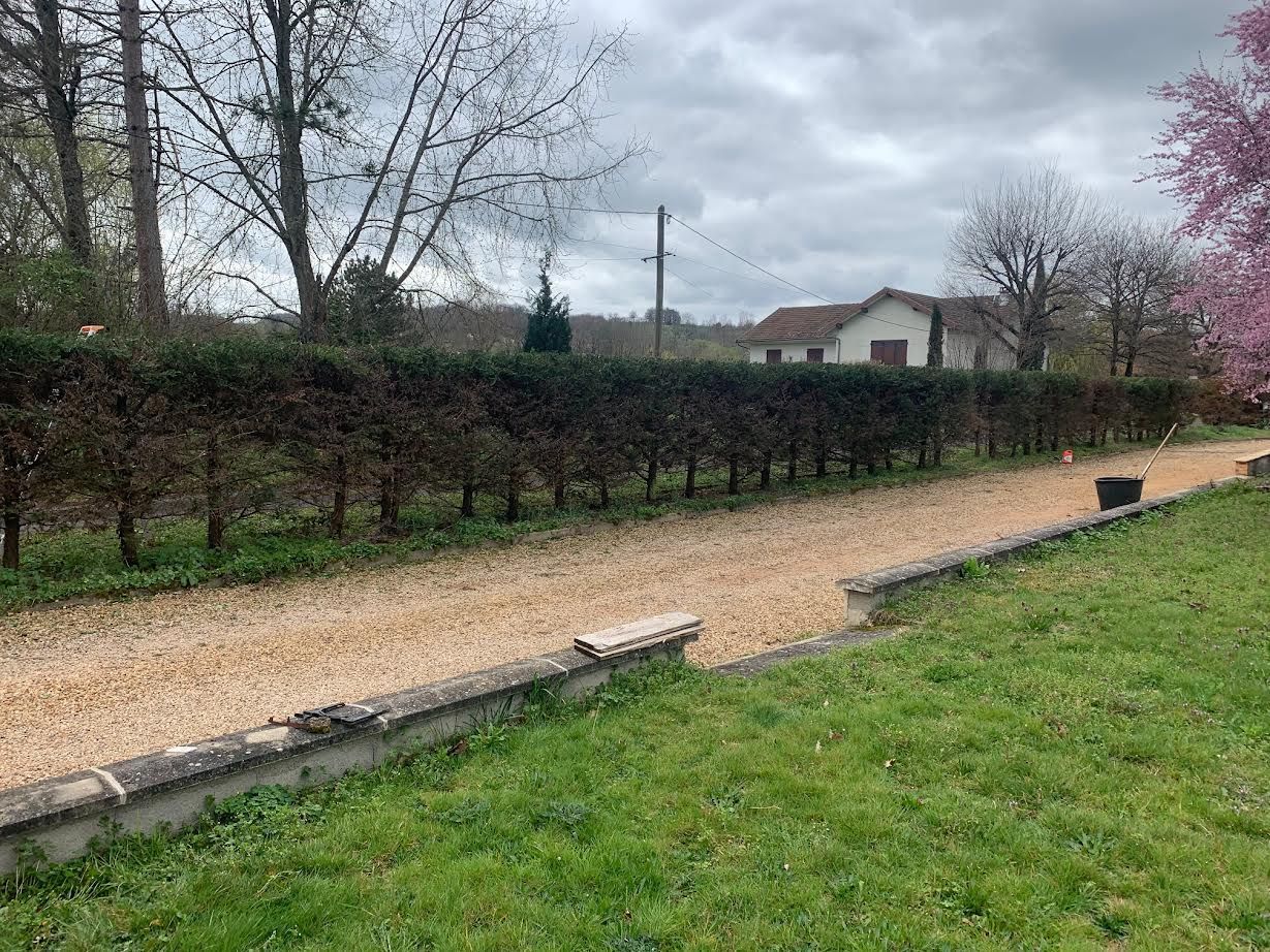 Chemin de gravier longeant une haie taillée et une pelouse, avec des arbres dénudés et une maison au loin sous un ciel nuageux.