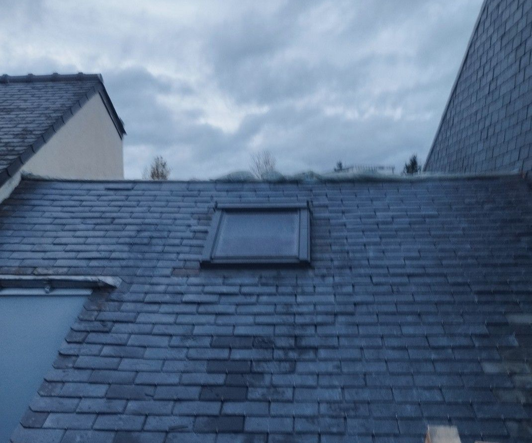 Toit en ardoise avec une lucarne rectangulaire sous un ciel nuageux.