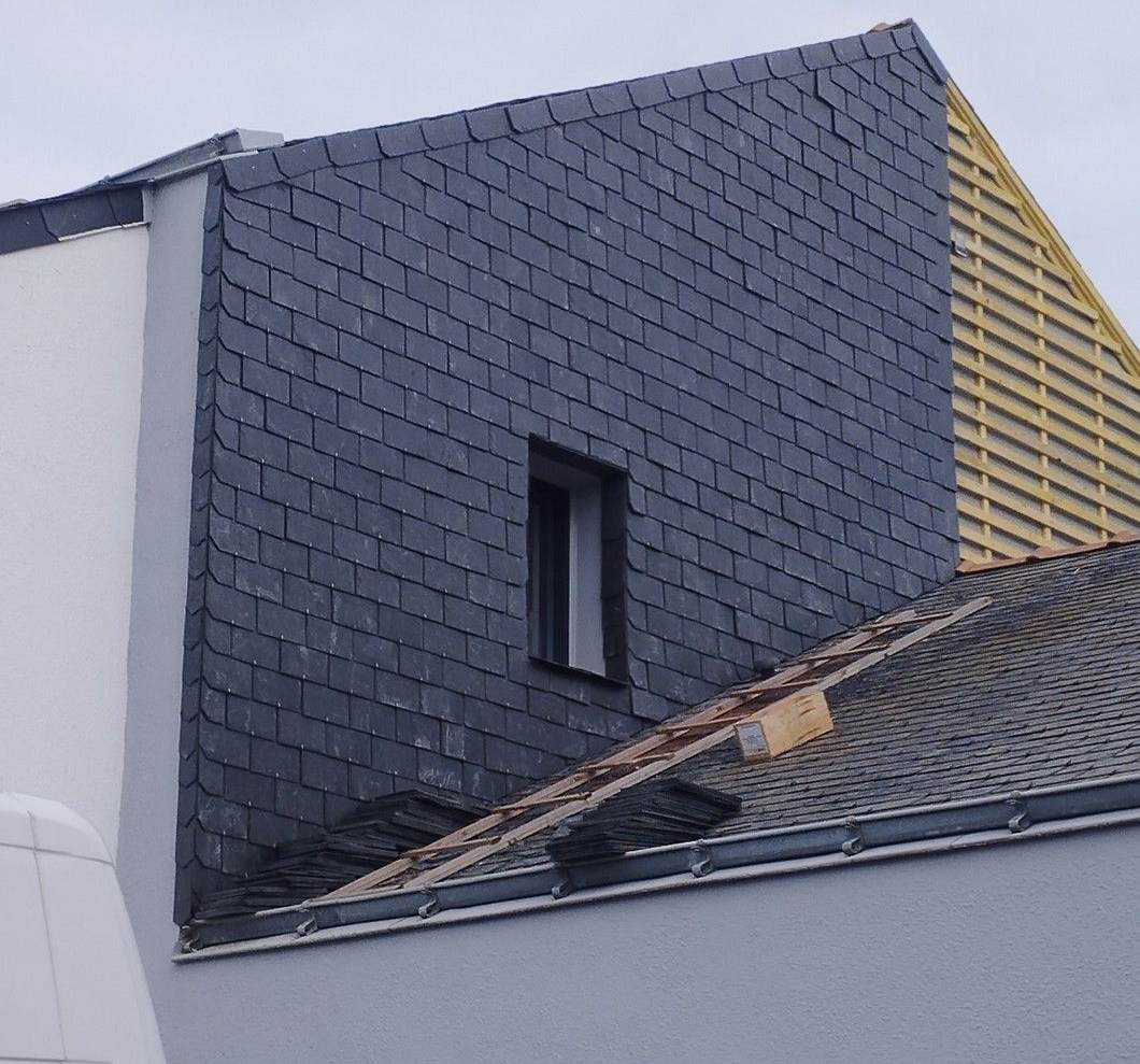 Façade d'immeuble en ardoise avec une petite fenêtre, toit en bois partiellement construit visible sur la droite.