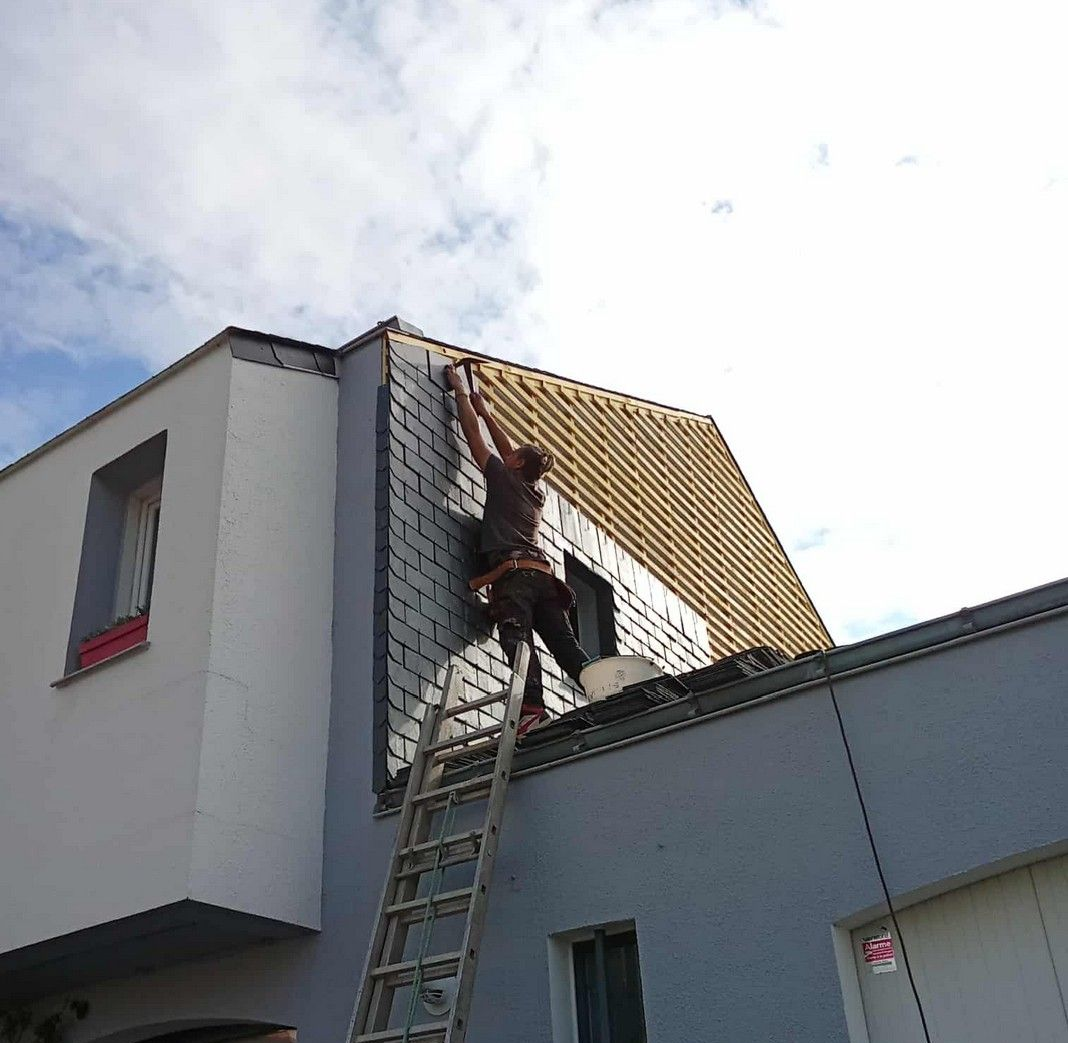 Un couvreur, juché sur une échelle, pose des ardoises gris foncé sur le toit d'une maison sous un ciel nuageux.