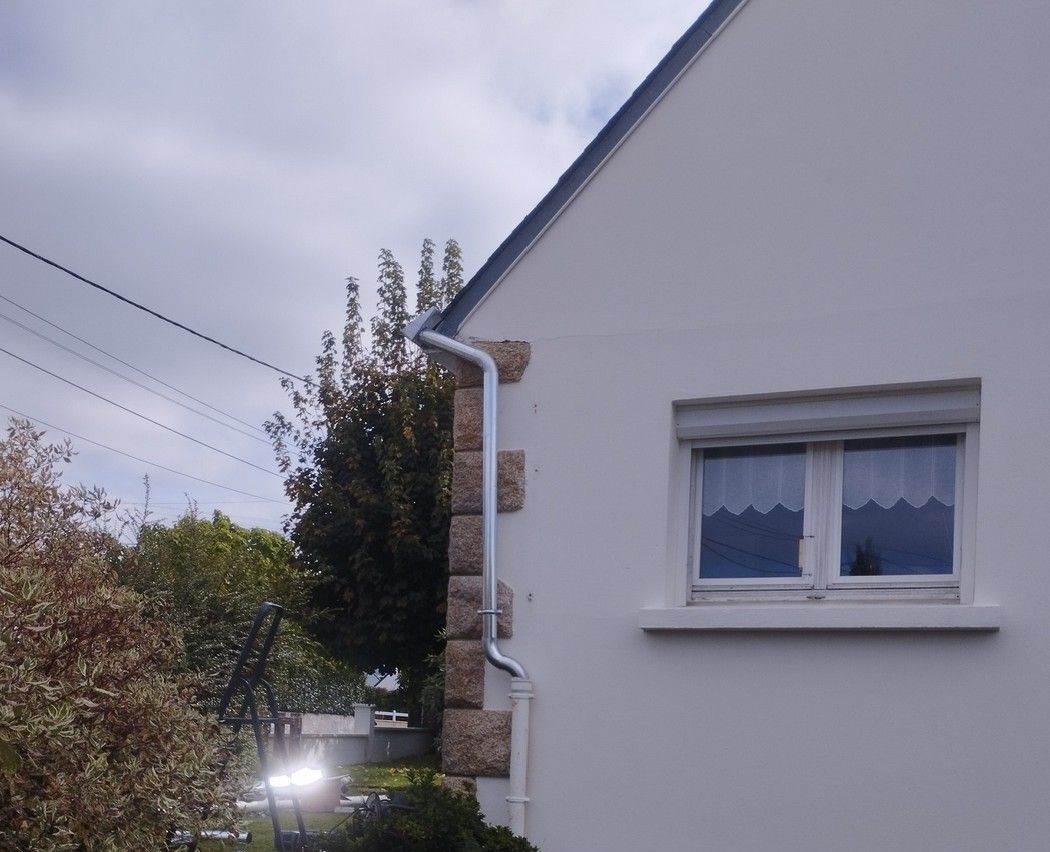 Coin d'une maison blanche avec un élément en pierre et une fenêtre, arbres et lignes électriques en arrière-plan.