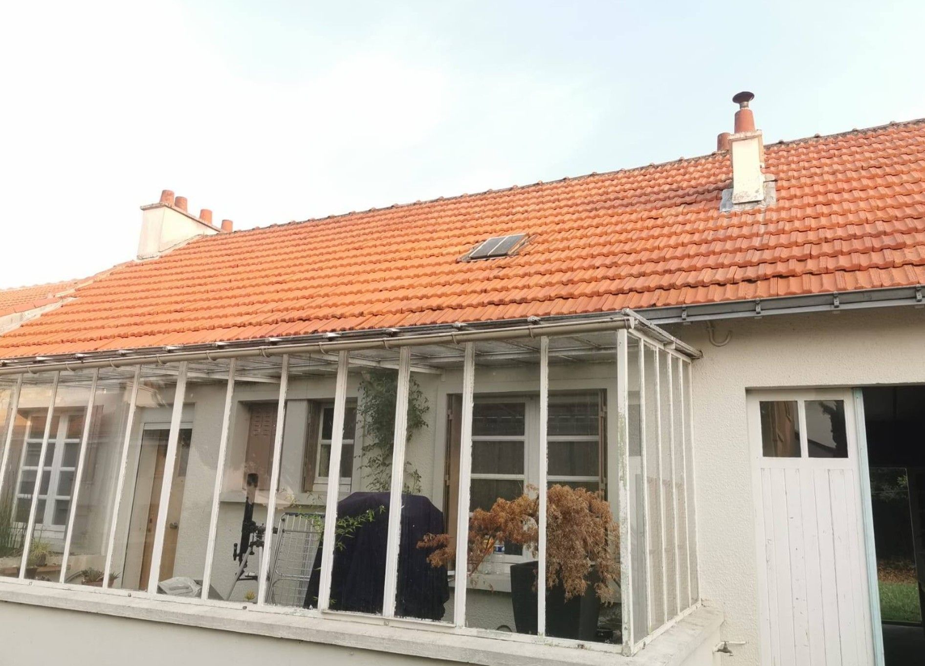 Maison aux murs blancs, avec une véranda vitrée et un toit de tuiles rouges, sous un ciel nuageux.