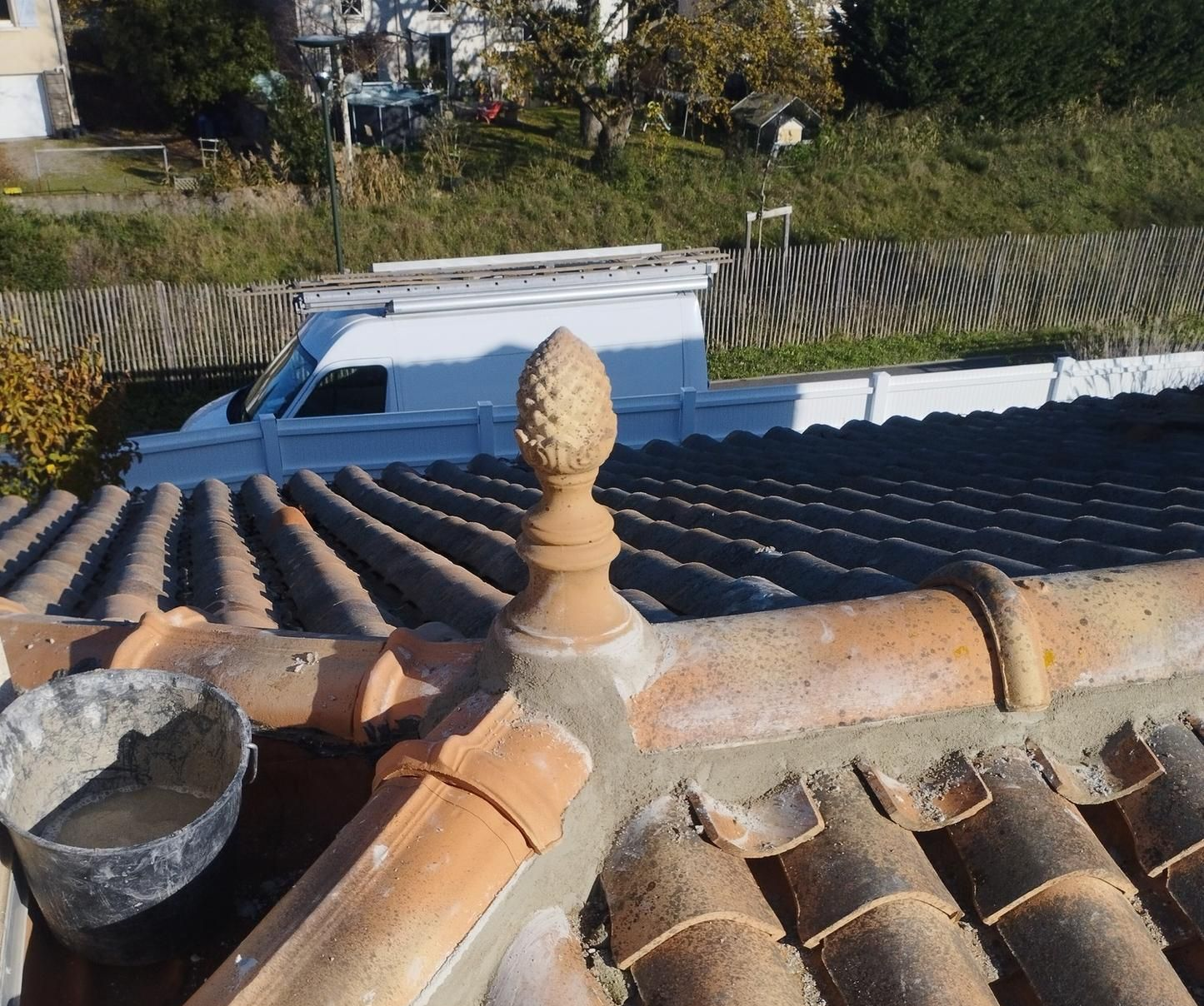 Toiture ornée d'un fleuron décoratif et de tuiles en terre cuite, camionnette blanche en arrière-plan.