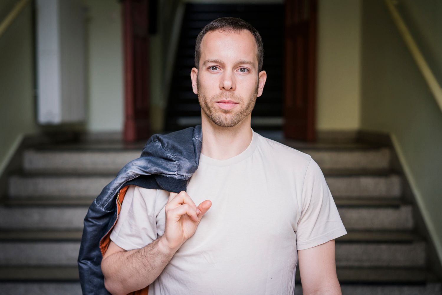 Ein Mann in weißem Hemd mit einer Lederjacke über der Schulter steht vor einer Treppe und schaut in die Kamera.