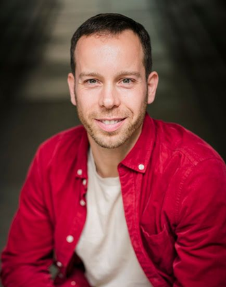 Mann lächelt; er trägt ein rotes Hemd über einem weißen Hemd. Grauer Hintergrund.