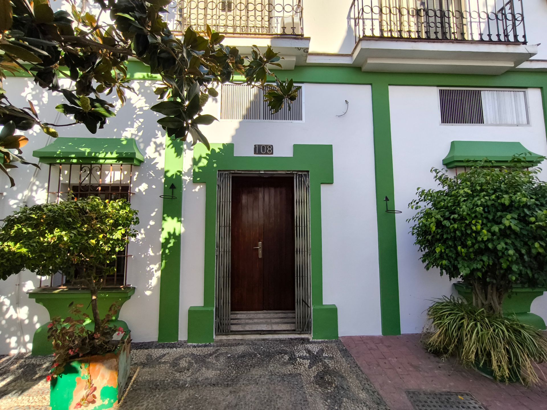 Edificio blanco con detalles verdes en un patio adoquinado con plantas en macetas y un cielo azul.